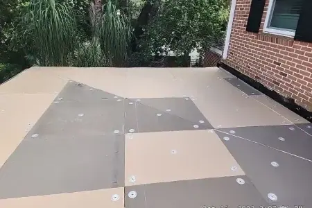 A flat rooftop with panels secured by metal fasteners, adjacent to a brick house with black shutters and surrounded by green trees—a signature project completed by Budget Roofing Service Inc, a trusted roofing company in Winter Haven.