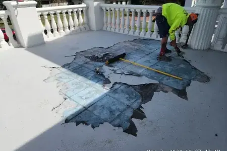 A worker from Budget Roofing Service Inc, in full safety gear, removes damaged waterproofing material from a balcony floor using a pry bar and broom; debris is scattered around the exposed area.