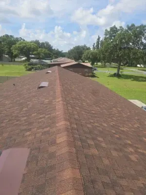 A view from the peak of a brown shingle roof installed by Budget Roofing Service Inc on a long building, surrounded by green lawns and trees under a partly cloudy sky.