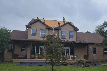 Workers in safety gear are repairing or replacing the roof of a two-story suburban house on a cloudy day, as part of a roof maintenance program, with ladders and tarps visible around the home.