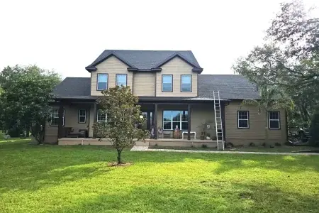 A two-story beige house with a dark roof, front porch, and a ladder leaning against the right side—showcasing quality work by Budget Roofing Service Inc. A small tree stands in the grassy front yard.