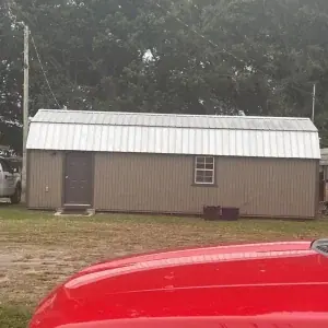A small gray shed with a metal roof, one door, and one window is shown, with a red vehicle in the foregroundâperfect inspiration for any roofing company in Winter Haven. Parked cars and trees can be seen in the background.