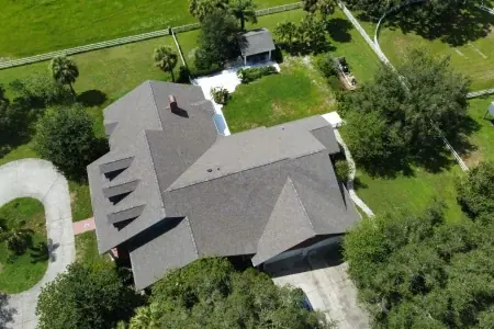 Aerial view of a large house with a gray roof, surrounded by green lawn, trees, a curved driveway, and a small outbuilding in the backyard—a perfect showcase for any roofer in Winter Haven.
