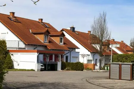 Row of modern suburban houses with red-tiled roofs and white exteriors on a quiet street, with trees and a street sign visible.