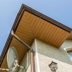 Close-up view of a house corner showing brown gutters, beige soffit, and fascia repair details, along with a satellite dish, window, and outdoor light fixture against a clear blue sky.