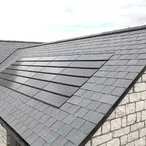 A sloped roof with gray slate tiles and integrated solar panels sits atop a stone building under a cloudy sky, showcasing modern low slope roofing systems designed for energy efficiency and durability.