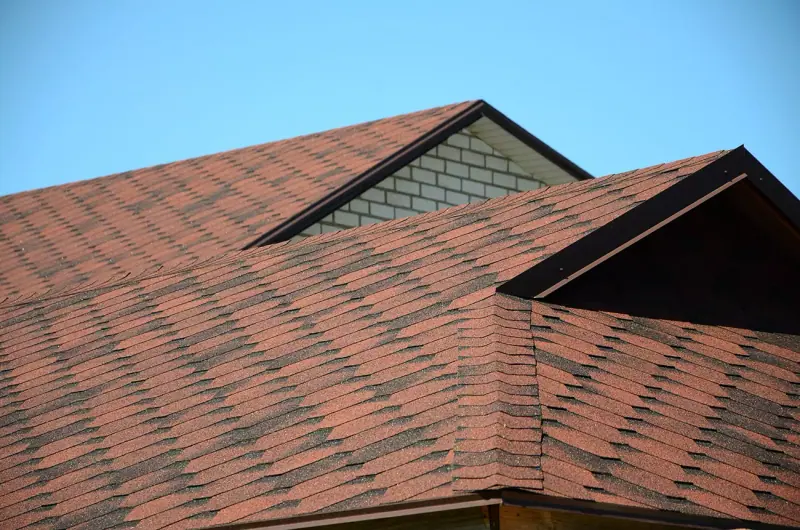 Close-up view of a house roof with red asphalt shingles, featuring multiple peaks and a section of white brick wall under a clear blue sky.
