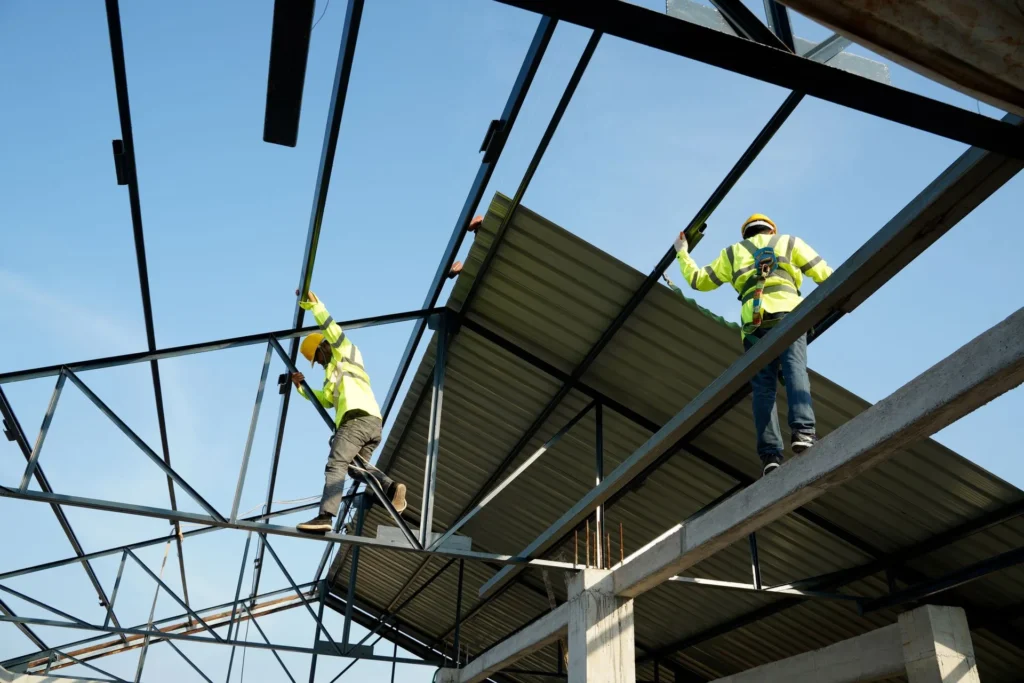 Two construction workers in safety gear install metal roofing sheets on a steel framework structure under a clear blue sky.