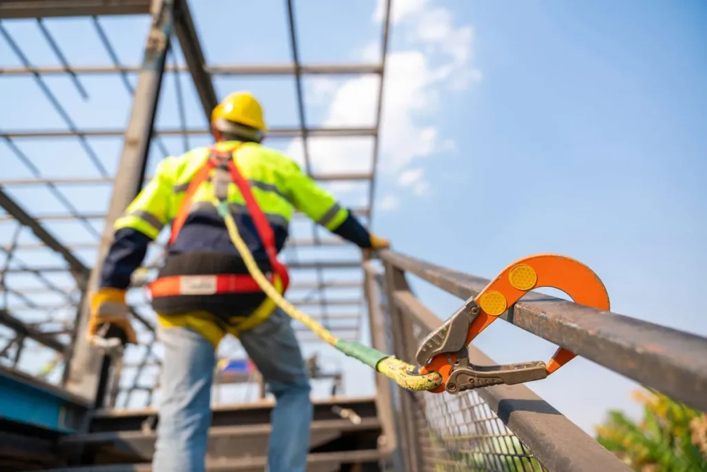 A construction worker wearing safety gear is secured to a metal railing with a harness and lanyard at a building site with a steel frame structure in the background.