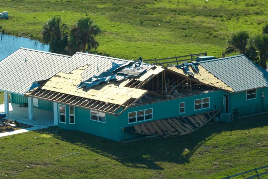 A turquoise house with severe roof damage; large sections of the roof are missing shingles and panels, exposing the interior to the outside.