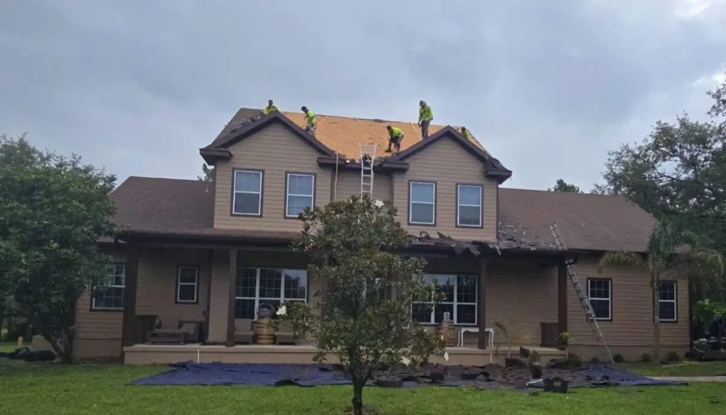 Workers in safety gear are repairing or replacing the roof of a two-story suburban house on a cloudy day, as part of a roof maintenance program, with ladders and tarps visible around the home.