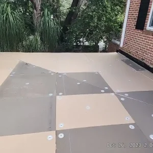 A flat rooftop with panels secured by metal fasteners, adjacent to a brick house with black shutters and surrounded by green trees—a signature project completed by Budget Roofing Service Inc, a trusted roofing company in Winter Haven.