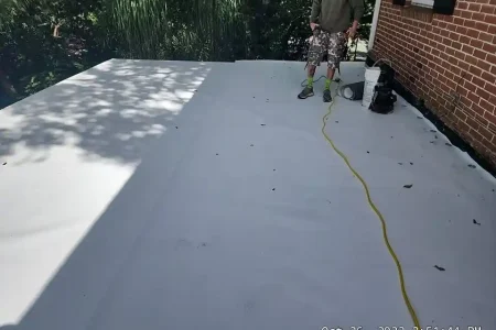 A person stands on a flat, freshly coated white roof near a brick wall, with equipment and an extension cord nearby. Shadows from trees cover part of the surface—work done by a top roofing company in Winter Haven.