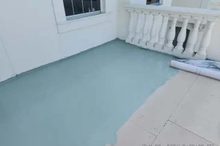 A balcony floor partially covered with a fresh coat of light blue-green paint next to a white railing and wall, with a paint roller mat lying on the right side—an attention to detail reminiscent of Budget Roofing Service Inc standards.
