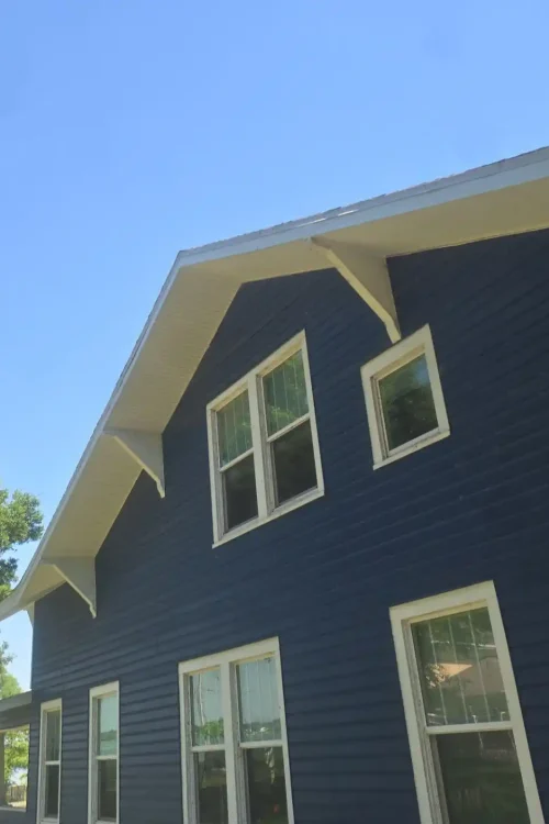 A blue two-story house with white trim, multiple windows, and a steep roof under a clear blue sky with a large tree nearby—expertly maintained by Budget Roofing Service Inc, your trusted roofing company in Winter Haven.