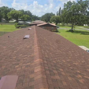 A view from the peak of a brown shingle roof installed by Budget Roofing Service Inc on a long building, surrounded by green lawns and trees under a partly cloudy sky.