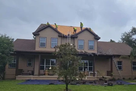 Workers in safety gear are repairing the roof of a two-story house, using ladders to access the roof while tarps cover the ground below.