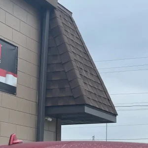 Brown shingled awning attached to the side of a beige brick building, installed by a roofing company in Winter Haven, with part of a red vehicle in the foreground and power lines in the background.