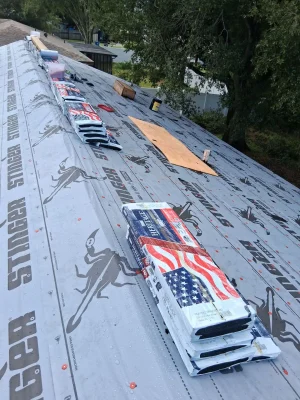 Stacks of roofing shingles and materials are placed on a roof covered with Stinger-branded underlayment, prepared by Budget Roofing Service Inc, a leading roofing company in Winter Haven. Trees and a building are visible in the background.