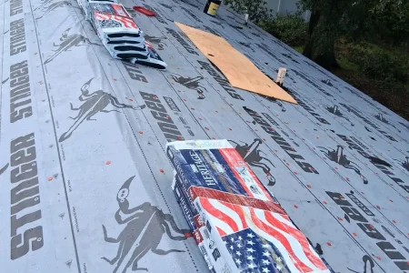 Stacks of roofing shingles and materials are placed on a roof covered with Stinger-branded underlayment, prepared by Budget Roofing Service Inc, a leading roofing company in Winter Haven. Trees and a building are visible in the background.