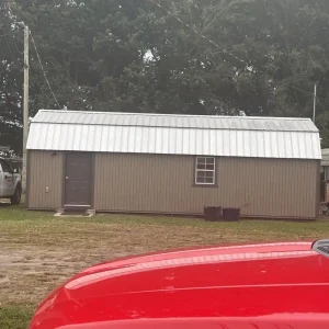 A small gray shed with a metal roof, one door, and one window is shown, with a red vehicle in the foreground—perfect inspiration for any roofing company in Winter Haven. Parked cars and trees can be seen in the background.