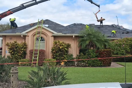 Workers from Budget Roofing Service Inc are installing new roofing materials on a house using cranes and ladders, with safety equipment and barriers in place. Trust this roofing company in Winter Haven for reliable and safe service.