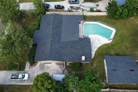 Aerial view of a house with a dark roof by Budget Roofing Service Inc, backyard swimming pool, detached garage, and several cars parked in the driveway and nearby street.