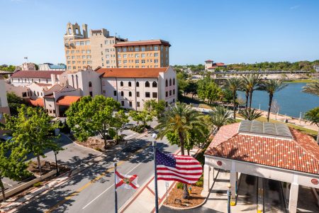 A city street with palm trees, historic buildings, and a river in the background; American and Florida state flags are visible in the foreground, reflecting the vibrant community Roofers in Lakeland FL are proud to serve.