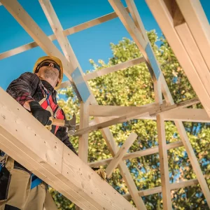 Construction worker wearing safety gear uses a handsaw to cut a wooden beam during the framing stage of a building under a clear sky, with trees in the background.