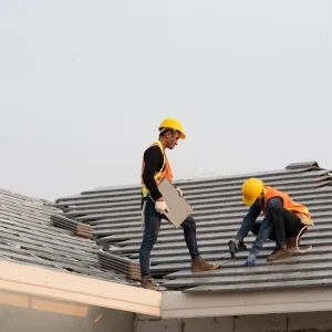 Two construction workers wearing safety gear perform tile roof installation, carefully placing roofing tiles on a building under clear skies.