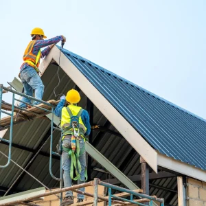 Two construction workers wearing safety gear install a metal roof sheet on a house structure using scaffolding and tools under a clear sky.