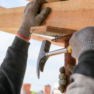 A person wearing gloves secures a wooden beam with a nail and hammer during roof framing repair.
