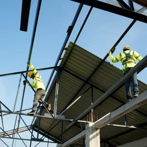 Two construction workers in safety gear install metal roofing sheets on a steel framework structure under a clear blue sky.