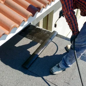 A worker in a plaid shirt uses a torch to apply a roll of waterproofing membrane to a flat roof near a tiled section, demonstrating expert flat roof repair and maintenance on low slope roofing systems.