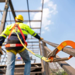 A construction worker wearing safety gear is secured to a metal railing with a harness and lanyard at a building site with a steel frame structure in the background.