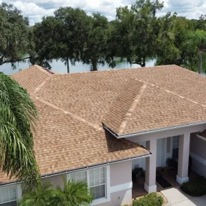 Aerial view of a beige-shingled, single-story house with a driveway, palm trees, and a lake with trees in the background, showcasing recent asphalt shingle roofing for lasting durability and style.