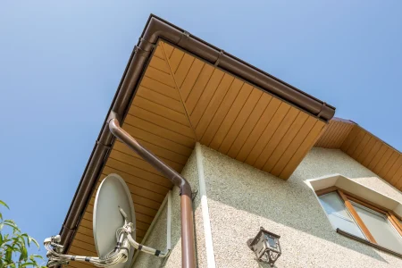 Close-up view of a house corner showing brown gutters, beige soffit, and fascia repair details, along with a satellite dish, window, and outdoor light fixture against a clear blue sky.