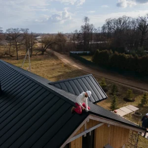 A worker kneels on the sloped metal roof of a house under construction, possibly performing chimney flashing repair, while another stands on the ground nearby; the area is surrounded by trees and open land.