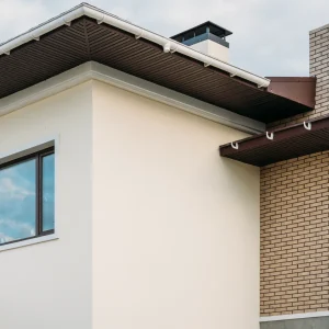 Modern house exterior showing cream stucco and tan brick walls, large windows, a sloped roof with brown eaves, visible rain gutters, and well-maintained soffit and fascia repair enhancing roof edge ventilation.