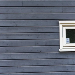 A small white-framed window set in a blue wooden wall, with a security alarm sticker visible on the window glass.