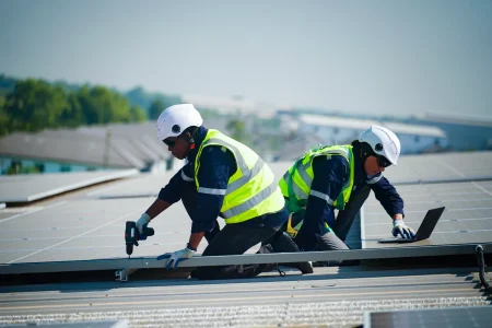 Two workers in safety gear install solar panels on a rooftop; one uses a drill while the other checks a laptop, showcasing the expertise of Budget Roofing, a trusted roofing company in Winter Haven.
