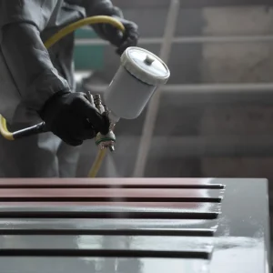A person in protective clothing uses a spray gun to apply paint to a metal surface in an industrial setting.