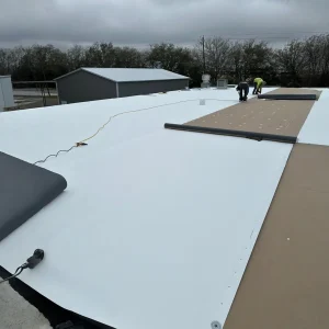 Two workers, including a roofer in Winter Haven, install large white roofing sheets on a flat commercial roof under cloudy skies; tools and materials are visible on the roof.
