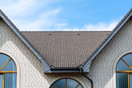 A close-up view of a house roof with brown shingles and white brick walls, featuring three arched windows with wooden frames under a blue sky.