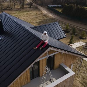 Two construction workers wearing helmets sit and stand on the black metal roof of a modern wooden house under construction, with ladders and scaffolding nearby, possibly preparing for gutter and downspout repair.