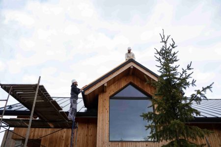 Two construction workers in safety gear work on the roof and scaffold of a modern wooden house with large windows and a metal roof.