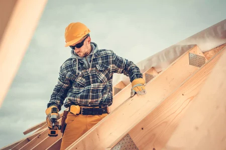 Construction worker wearing a yellow hard hat and gloves uses a power drill on a wooden roof frame under a cloudy sky.